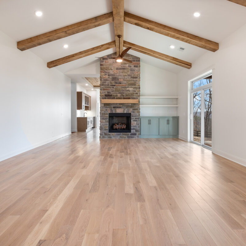 photo of a large living room with wood floors, white walls, stone fireplace with custom wood mantle, built-in shelving, and large rustic wooden beams on ceiling